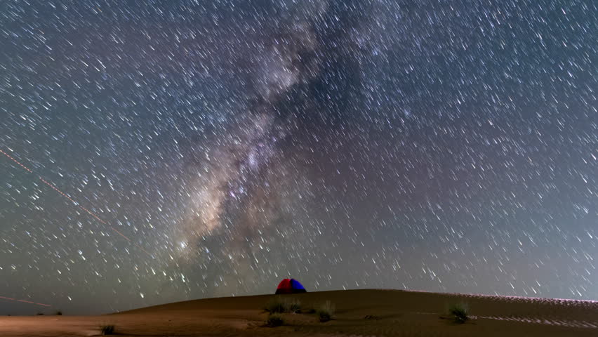 Stunning 4k timelapse of the Milky Way galaxy over a desert campsite with glowing tents near an oasis and date palm trees. Clear night sky, star scape, and sand dunes in a remote wilderness location. 