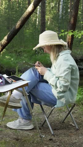 A romantic girl artist, inspired by the forest nature, paints a landscape by a forest river. Mosquitoes bite and do not give rest. The creative process. Vertical video, shorts.