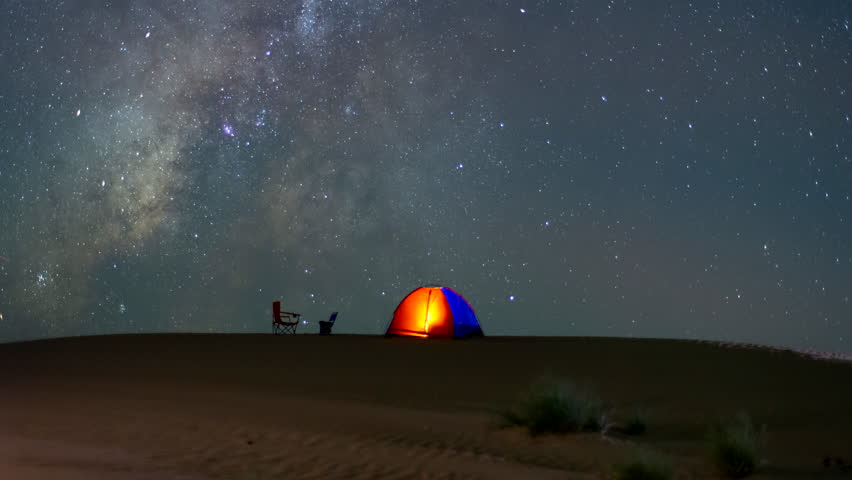Stunning 4k timelapse of the Milky Way galaxy over a desert campsite with glowing tents near an oasis and date palm trees. Clear night sky, star scape, and sand dunes in a remote wilderness location. 