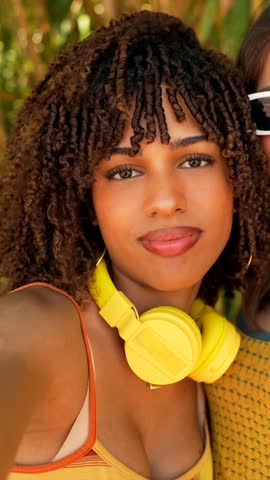 Young women wearing stylish white sunglasses and yellow headphones sharing joyful moments, smiling and posing together during summer leisure time outdoors