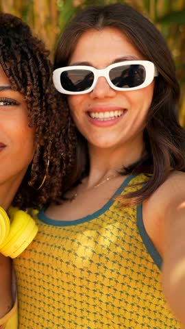 Young women wearing stylish white sunglasses and yellow headphones sharing joyful moments, smiling and posing together during summer leisure time outdoors
