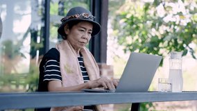 asian senior woman uses laptop at outdoor cafe during solo travel wearing stylish hat scarf black striped shirt working peacefully near water pitcher glass nature background - Powered by Shutterstock - Get 15% off with code: PIKWIZARD15
