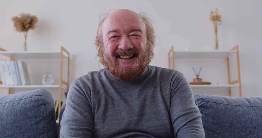 Happy elderly man sitting on sofa and looking at camera during online video call. Cheerful senior male engaged in conversation laughing and actively gesticulating while telling funny stories.
