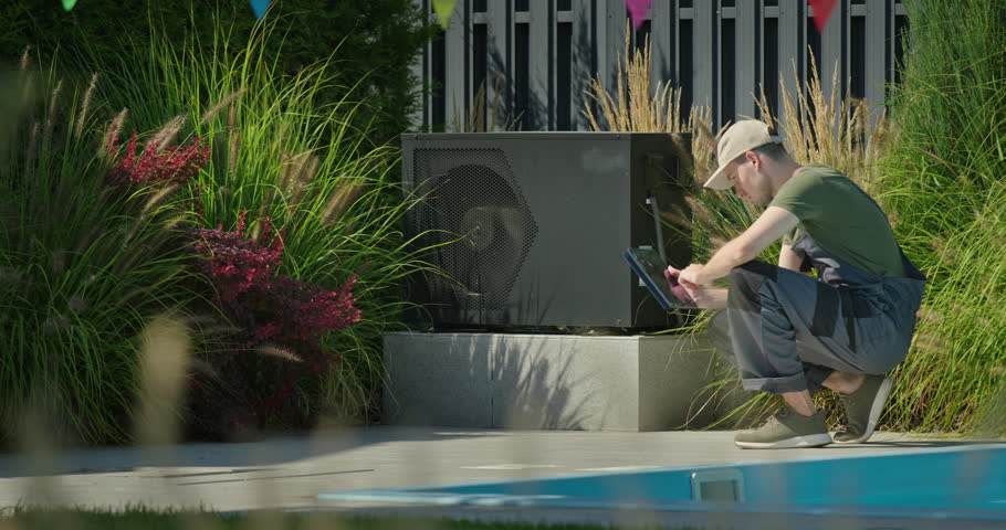 Young technician squatting near poolside heat pump with tablet, surrounded by plants and colorful flags, performing outdoor maintenance work. High quality 4k footage