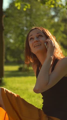 Happy hadsome female sitting on green grass in City Park and enjoying friendly conversation via digital smartphone device Vertical shot.