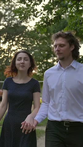 Front portrait view of a young couple, man and woman, holding hands walking along a path in a forest along a lake. people play volleyball at thebackground. Sunset. Vertical shot.