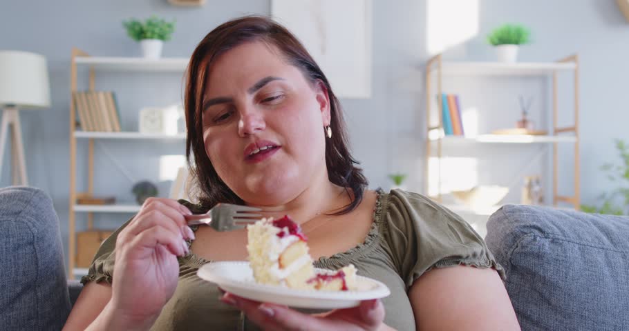 Portrait of happy smiling fat young overweight woman eating slice of tasty cake on plate enjoying sweet dessert sitting on sofa in living room at home. Obesity and dieting concept. 4k video.