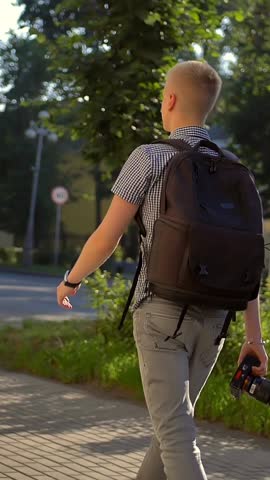 Back-side view of student photographer with a black backpack is walking along a town street. Expensive black digital camera in hands. He looks around, looking for interesting sights. Vertical shot.