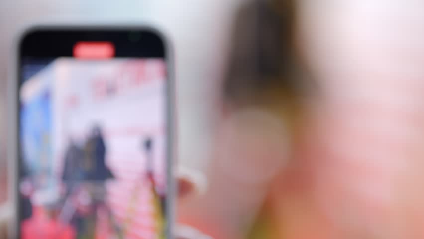Blurry image of a person holding a cell phone with a video recording app. The person appears to be taking a selfie, but the focus is on the blurry background and the person's face