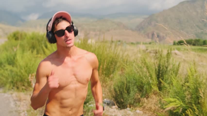 A muscular young man runs a mountain road under cloudy skies, singing along and moving to the beat of music in his headphones. The rhythm drives his pace, blending power and pure joy of running.