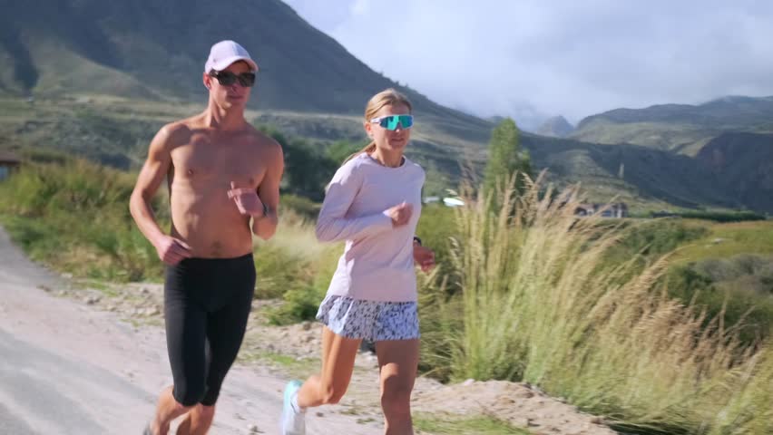 An athletic couple walks a sunny mountain road, checking smartwatch timers before starting interval training. As mist curls over distant peaks, they ease into a run to begin intense race preparation.