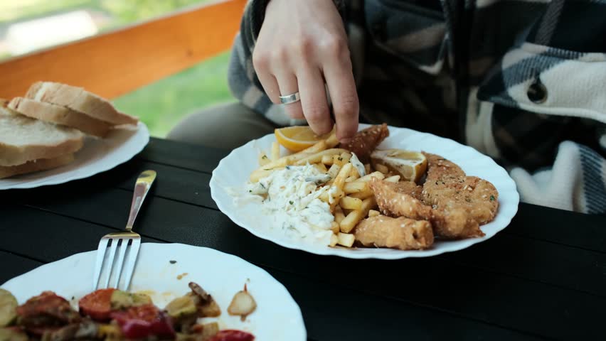 Young woman eats French fries with white sauce on a wooden cabin terrace. Horizontal 4k footage. Close view of girl with food