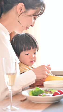 A Japanese woman and her toddler eating dinner in a dining room bathed in beautiful backlighting. A vertical video showing a close-up of the cute toddler and the mother feeding him.