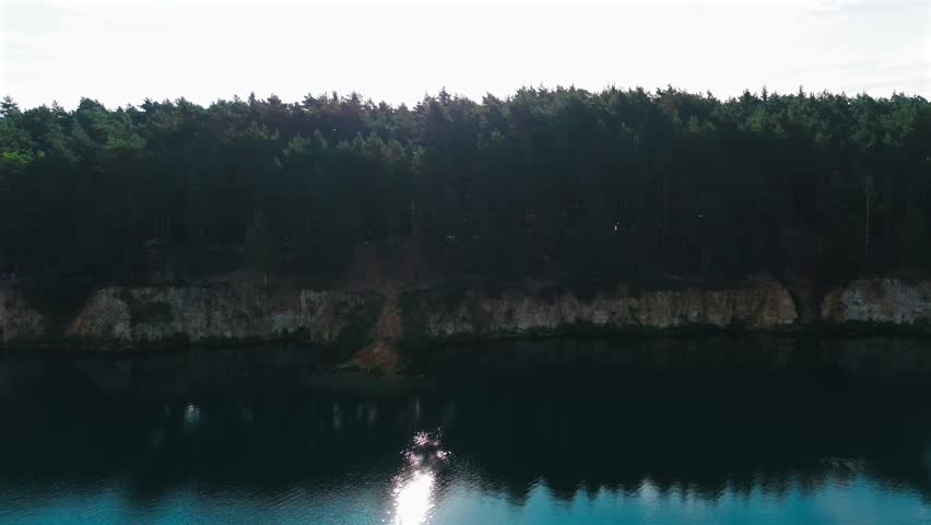 A drone captures a scenic view of a calm blue lake surrounded by steep chalk cliffs and dense pine forest under direct sunlight.