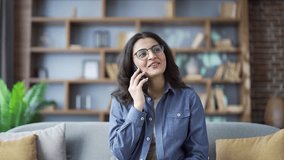 Happy young woman talking on smartphone while sitting on sofa in the living room at home. Friendly pretty female freelancer in glasses has a conversation with a client or communicates with a colleague - Powered by Shutterstock - Get 15% off with code: PIKWIZARD15