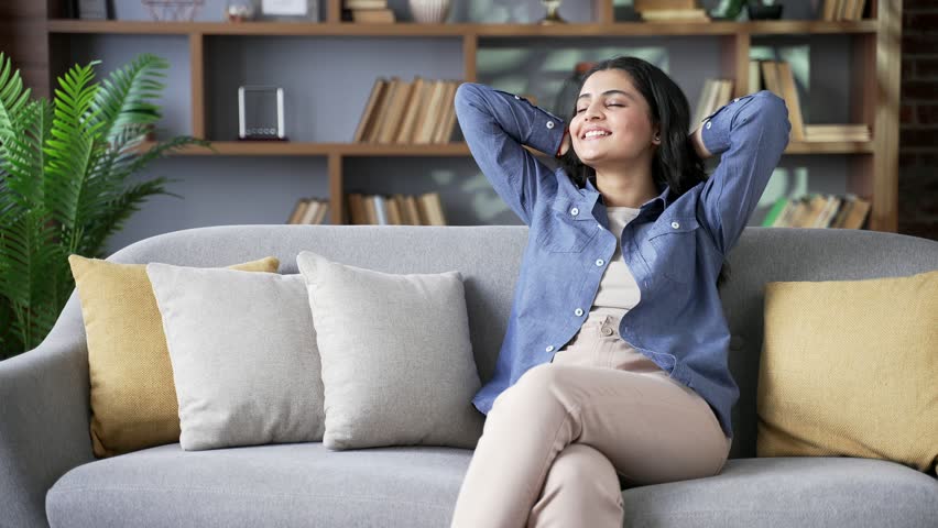 Young happy woman relaxing with her eyes closed and her hands behind her head while sitting on a comfortable sofa in the living room at home. Calm beautiful female resting at leisure, feeling relieved