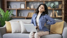 Tired young woman suffering from back pain while working on a laptop sitting on sofa in living room at home. Upset exhausted female massages painful lower back muscles. Discomfort due to poor posture - Powered by Shutterstock - Get 15% off with code: PIKWIZARD15