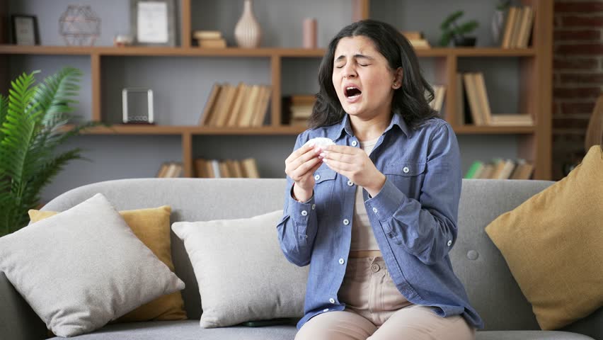 Allergic young woman sneezes and wipes her nose with handkerchief. Unhealthy adult female suffering from seasonal allergies sitting on sofa in living room at home. Girl is sick with cold, virus or flu