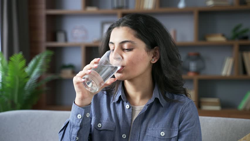 Young adult woman drinks water from a glass while sitting on the sofa in the living room at home. Happy smiling female feeling relieved, enjoying a clean cool drink, relaxing and resting. Close up