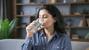 Young adult woman drinks water from a glass while sitting on the sofa in the living room at home. Happy smiling female feeling relieved, enjoying a clean cool drink, relaxing and resting. Close up - Powered by Shutterstock - Get 15% off with code: PIKWIZARD15