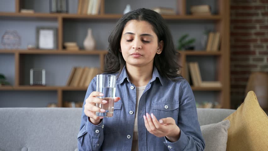 Young woman takes painkillers and drinks water from a glass while sitting on the sofa in the living room at home. Sick unhealthy adult female suffering from a cold, unwell, feeling bad. Close up