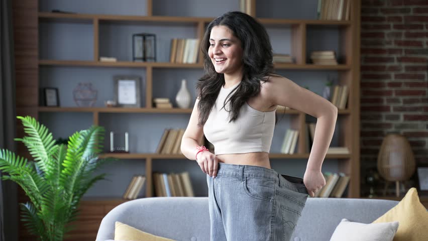 Happy young adult woman celebrating weight loss success at home wearing oversized pants smiling with joy and confidence in living room. She looks joyfully at camera with happiness inspiring on fitness