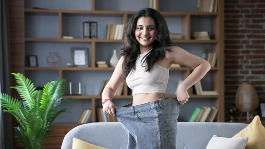 Happy young adult woman celebrating weight loss success at home wearing oversized pants smiling with joy and confidence in living room. She looks joyfully at camera with happiness inspiring on fitness