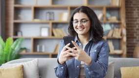 Young smiling woman in glasses using mobile phone sitting on sofa in living room at home. Happy beautiful female student browses social media, chats online, writes or reads messages on smartphone - Powered by Shutterstock - Get 15% off with code: PIKWIZARD15