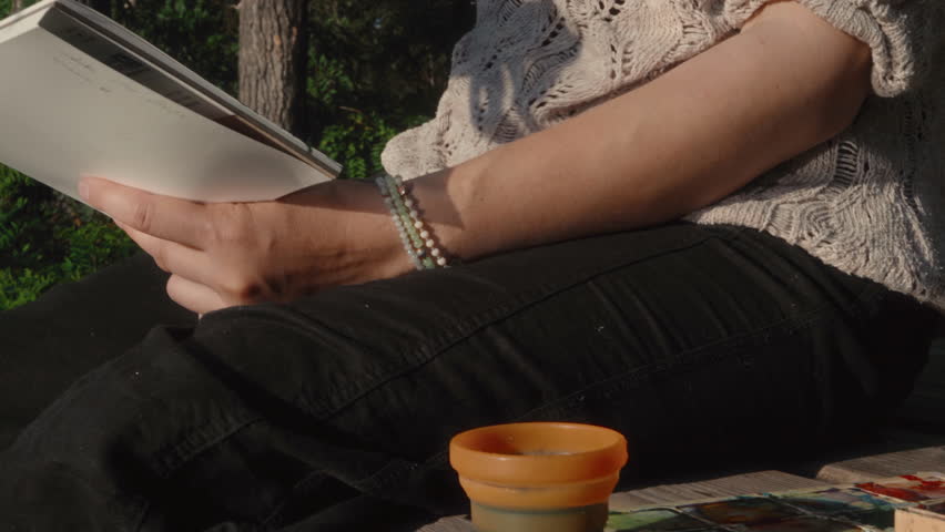 The hand of a female artist washes a paint brush in a glass of water on a wooden pier near a river