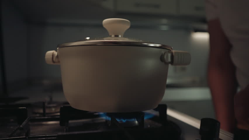 Close-up hands of chef male placing covered pot onto gas stove and turning burner, preparing for cooking session in dark professional kitchen environment. Shooting in slow motion.