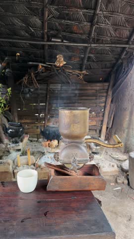 Authentic rustic scene: aged coffee grinder, steaming kettles, and a simple cup on a weathered wooden table within a traditional thatched hut. Evokes feelings of warmth, history, and handcrafted