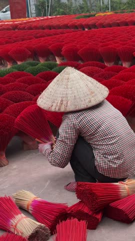 A person in a conical hat meticulously bundles vibrant red incense sticks, creating a striking contrast against the countless bundles surrounding them. The scene offers a glimpse into traditional
