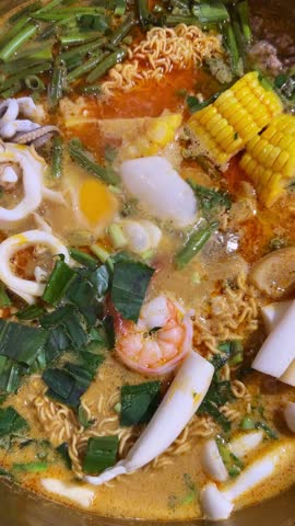 A person adds coconut milk to a delicious bowl of Tom Yum soup with noodles, shrimp, squid, and mushrooms. The vibrant colors and fresh ingredients make this a mouthwatering culinary experience.