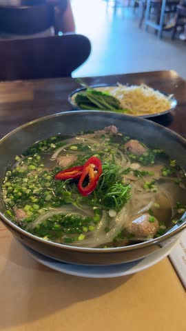 A steaming bowl of Pho, a traditional Vietnamese noodle soup, garnished with fresh herbs, chili peppers, and beef meatballs. The appetizing broth and vibrant ingredients create a delicious visual.
