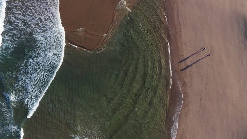 4K top-down view of couple walking along sandy beach washed by Atlantic Ocean waves in Portugal casting long shadows in low sunrise light. Shot during the Camino Portuguese —  popular pilgrimage route