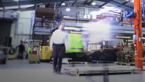 Time-lapse clip of busy workers in a warehouse or factory, wearing high visibility clothing and hard hats. They are checking stock levels and using a forklift truck to move empty wooden pallets.