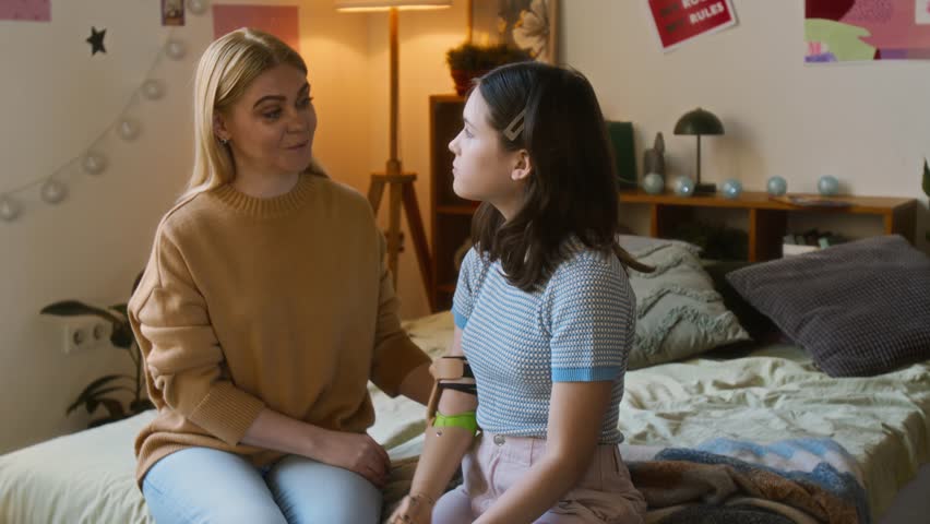 Middle-aged woman carefully helping her teenage daughter adjust her prosthetic arm while sitting together on bed in cozy room - Powered by Shutterstock - Get 15% off with code: PIKWIZARD15