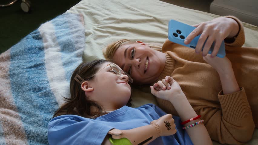 High angle shot of smiling mother and teenage daughter with prosthetic arm playing on smartphone and lying on bed - Powered by Shutterstock - Get 15% off with code: PIKWIZARD15