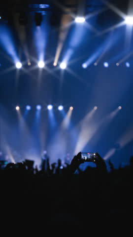 Concerthall show. Medium close-up shot. Vertical shot. Woman hands with phone in foreground with a clapping crowd of fans