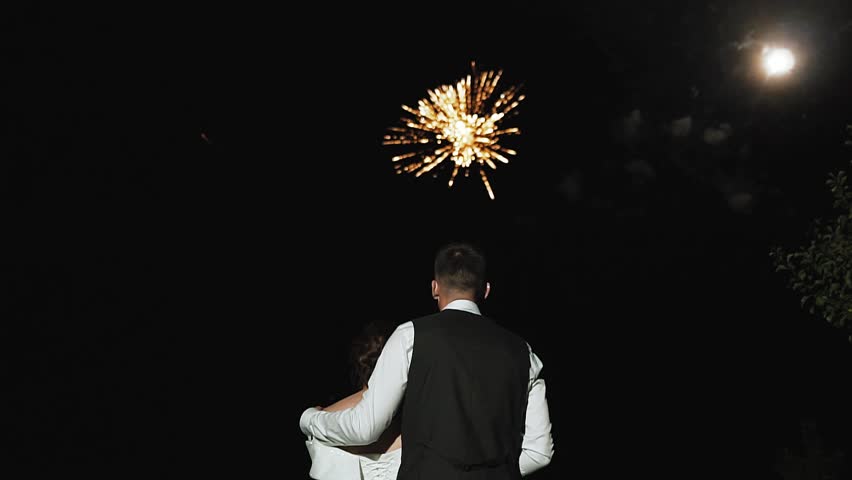 Unrecognizable newlywed couple hugging watching fireworks in night sky at a wedding. Gorgeous wedding fireworks, wedding day celebration. Sparks from the fireworks beautifully scatter in the dark sky.