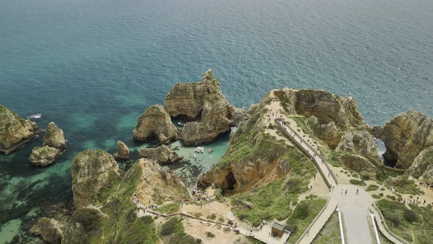 Drone orbits at high angle over Ponta da Piedade as tourists walking and in boats explore the caves and rock formations in Lagos, Portugal
