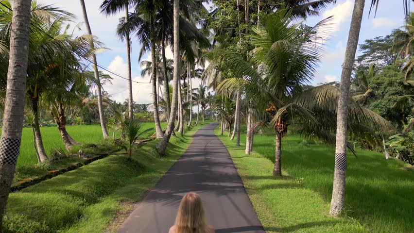 Cinematic shot of woman in red dress walking along scenic palm-lined road surrounded by rice fields in Bali. Peaceful tropical lifestyle and travel scene in Indonesia.
