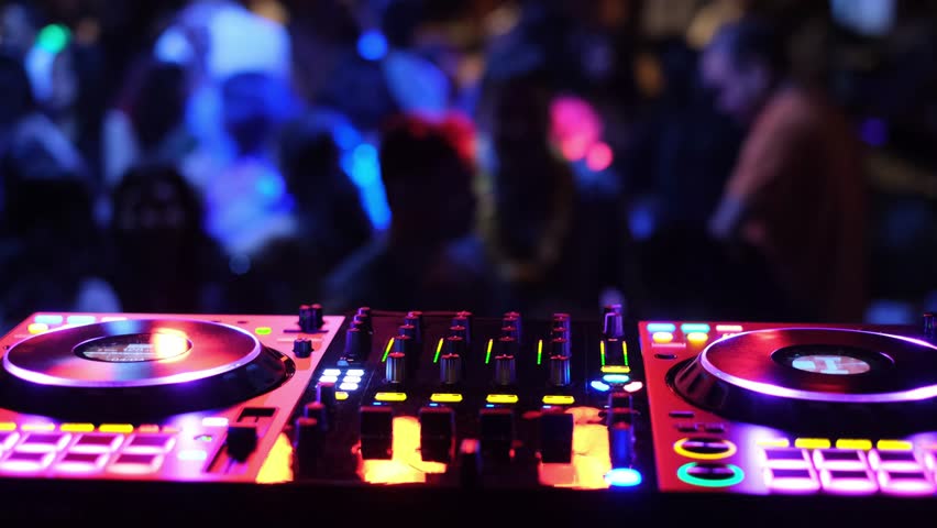 A DJ console that is vibrant and colorful, illuminated with vivid lights, at a lively party with a large dancing crowd