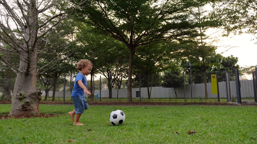 Barefoot young boy runs with soccer ball at dog park, tracking slow motion shot. Boy pushes ball ahead and runs, then aims and kicks toward fence. Training on green grass at enclosed dog run area.