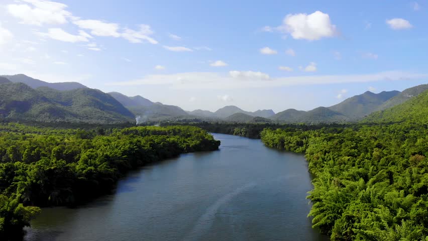 Ratchaburi Thailand. 20 June 2024, An expansive aerial or wide shot of a serene river winding through lush green mountains and dense forests under a bright blue sky with scattered clouds.