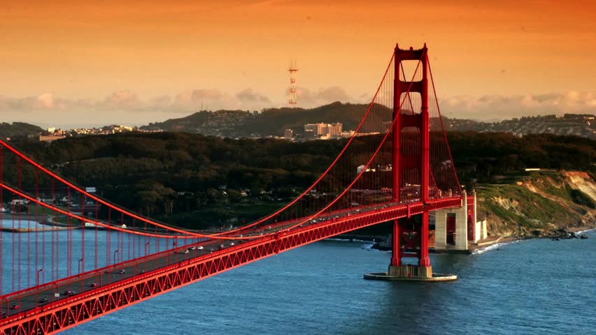 Golden Gate Bridge illuminated by sunset, casting warm hues over San Francisco Bay. Cars travel across bridge while gentle waves reflect colorful sky. Perfect backdrop for urban scenery.