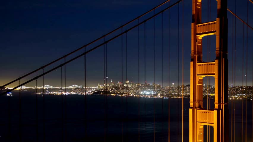 Illuminated Golden Gate Bridge showcases its vibrant orange color against a night sky. San Francisco skyline sparkles with city lights. Stars twinkle above calm waters.