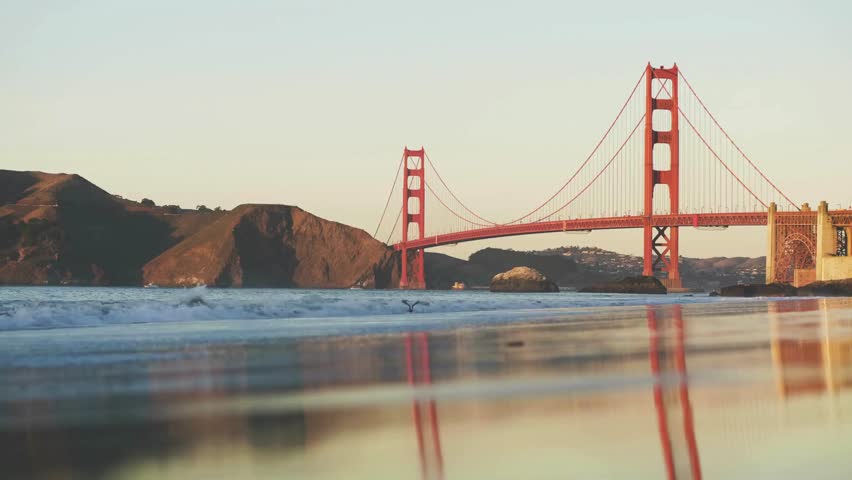 Witness the stunning sunset as it casts a warm glow over the famous bridge. Waves crash along the shore, creating a serene backdrop against the vibrant evening sky, capturing nature