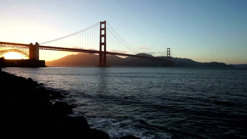 Sunset view of Golden Gate Bridge shining over San Francisco Bay. Calm waters reflect warm hues while mountains rise in background, showcasing California