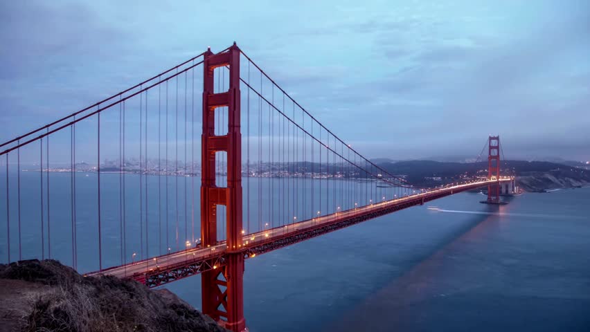 Golden Gate Bridge stands tall against dusk sky, twinkling lights reflecting in water below. San Francisco